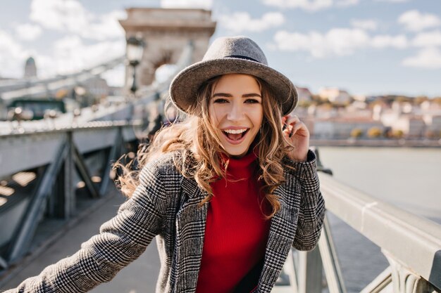 Encantadora chica rizada en elegante sombrero gris posando en el puente en un día soleado