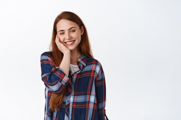 Encantadora chica romántica con pelo largo jengibre, ruborizada y sonriente, encantada de pie en blanco