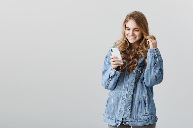 Encantadora chica rubia coqueta sonriendo al smartphone intrigado