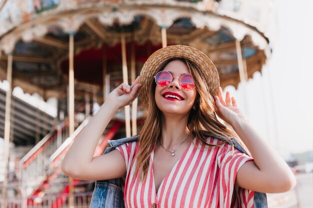 Encantadora chica rubia tocando su sombrero de paja y riendo en verano. Foto al aire libre de alegre mujer europea posando delante del carrusel.