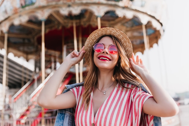 Encantadora chica rubia tocando su sombrero de paja y riendo en verano. Foto al aire libre de alegre mujer europea posando delante del carrusel.