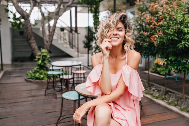 Encantadora chica rubia en vestido romántico sentado en la cafetería de la calle. Mujer joven entusiasta posando en restaurante con plantas