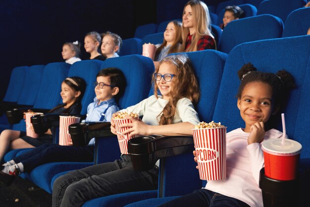 Encantadora chica sentada en el cine con amigos, mirando a cámara y sonriendo mientras ve una película. Adorable niña africana comiendo palomitas de maíz y bebiendo agua dulce