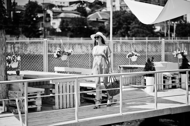 Encantadora chica con sombrero transparente y gafas de sol caminando por el muelle junto al lago con su bolso Foto en blanco y negro