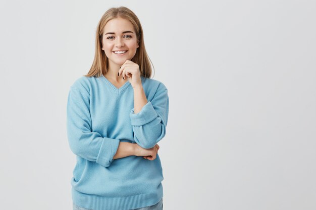 Encantadora chica vestida de azul sonriendo con dientes blancos, sosteniendo la mano debajo de la barbilla, pensando en planes para vacaciones. Mujer con cabello rubio mirando con expresión complacida y tranquila.