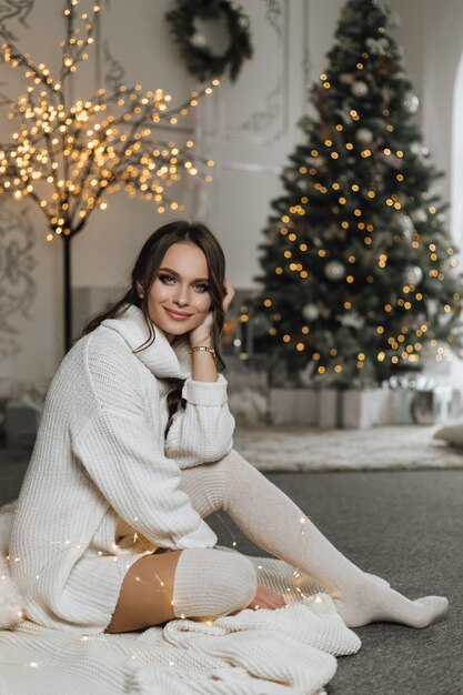 Encantadora chica con un vestido de punto tiene un aspecto de ensueño y se sienta sobre un fondo de árbol de Navidad