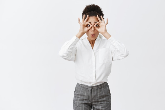 Encantadora compañera de trabajo despreocupada e infantil con camisa blanca y pantalones haciendo círculos con las manos sobre los ojos, doblando los labios como si mirara a través de binoculares algo asombroso y asombroso