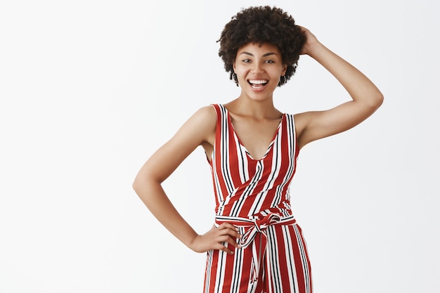 Foto gratuita encantadora y coqueta mujer afroamericana hermosa con peinado rizado en un mono a rayas de moda sosteniendo la mano detrás de la cabeza y el brazo en la cintura, sonriendo con alegría posando sobre una pared gris