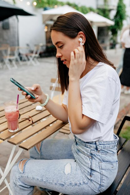 Encantadora dama adorable con camiseta blanca y jeans tocando auriculares y escuchando música al aire libre en la terraza de verano Linda mujer Lin auriculares inalámbricos en la terraza de verano