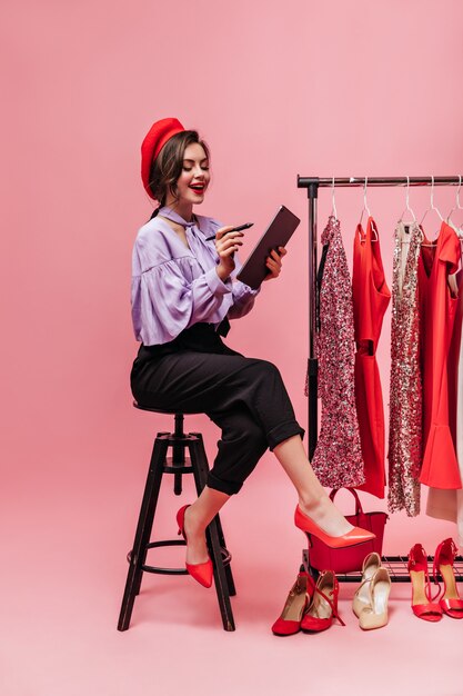 Encantadora dama en blusa y pantalones de moda toma notas en tableta. Chica posando sobre fondo de vestidos.