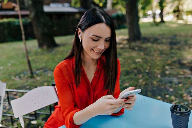 Encantadora dama bonita con camisa roja sentada en un café al aire libre con el teléfono inteligente y escuchando música