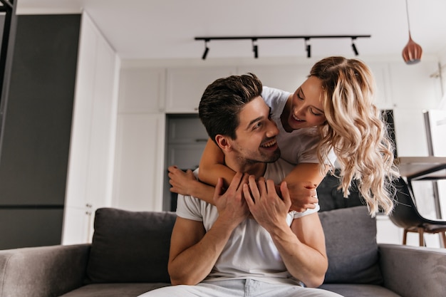 Encantadora dama caucásica abrazando a su marido con amor. Retrato de interior de increíble pareja divirtiéndose en fin de semana.