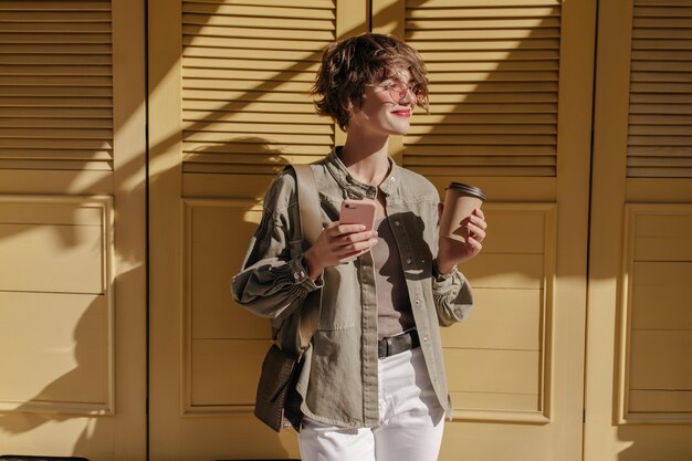 Encantadora dama con chaqueta verde oliva y pantalones blancos sostiene una taza de té y un teléfono sobre un fondo brillante Chica morena con gafas sonríe sobre un fondo amarillo