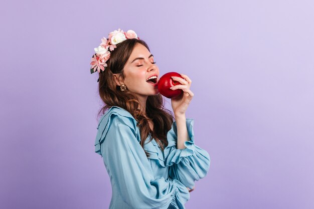 Encantadora dama en una corona de flores muerde jugosa manzana. Retrato de niña de blusa azul en la pared lila.