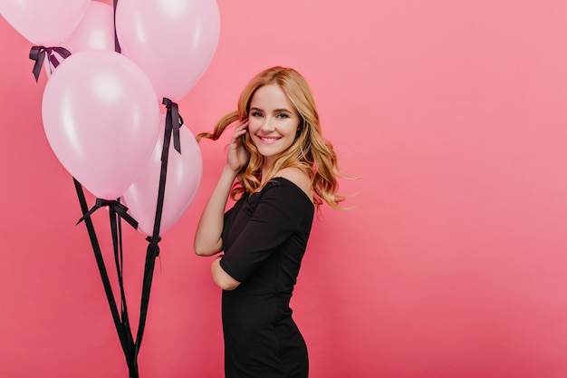 Encantadora dama delgada jugando con su cabello rubio mientras está de pie cerca de globos. Foto interior de encantadora niña feliz cumpleaños relajante durante la fiesta.