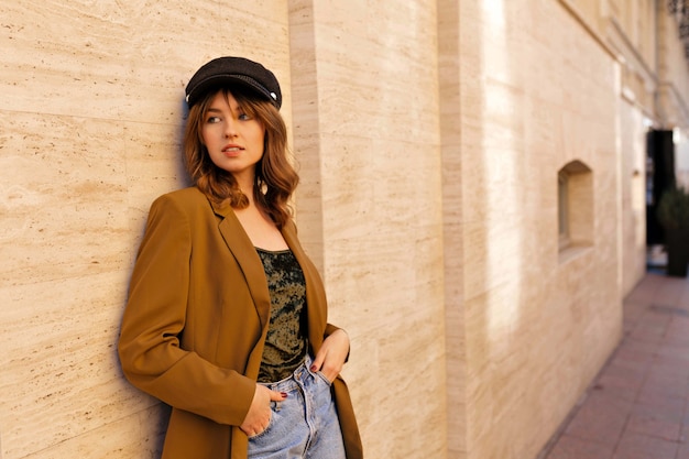 Encantadora dama elegante en chaqueta mostaza de moda y gorra negra posando afuera en un día cálido y soleado