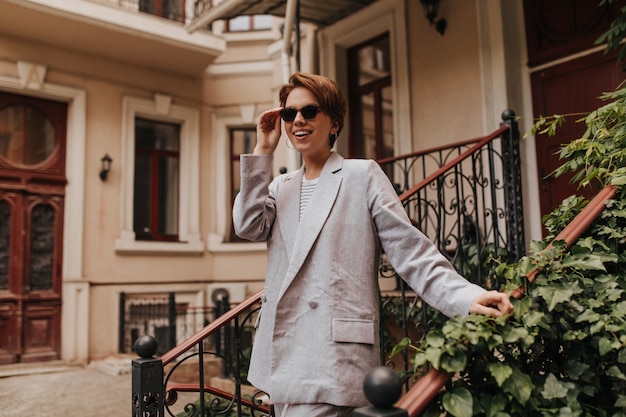 Encantadora dama en elegante traje se quita las gafas de sol y camina afuera. Mujer joven en chaqueta gris y pantalones sonriendo frente al edificio