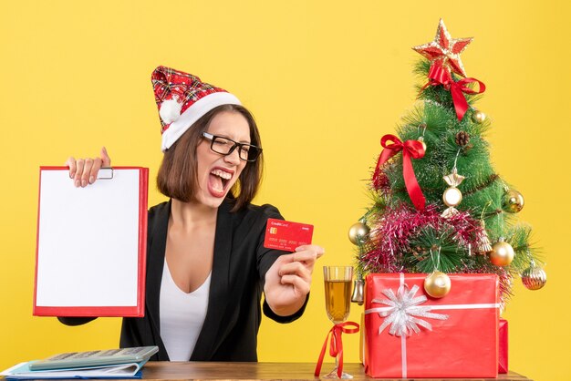 Encantadora dama emocional en traje con sombrero de santa claus y anteojos mostrando tarjeta bancaria y documento en la oficina en amarillo aislado