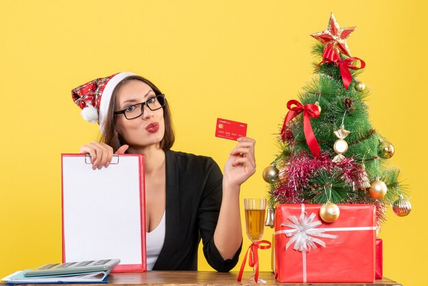 Encantadora dama emocional en traje con sombrero de santa claus y anteojos mostrando tarjeta bancaria y documento en la oficina en amarillo aislado