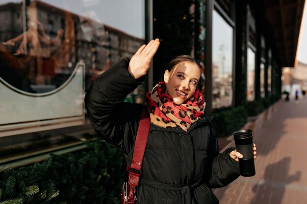 Encantadora dama encantadora con una sonrisa maravillosa con una chaqueta oscura y una bufanda roja está bebiendo café y cubriendo su mano del sol