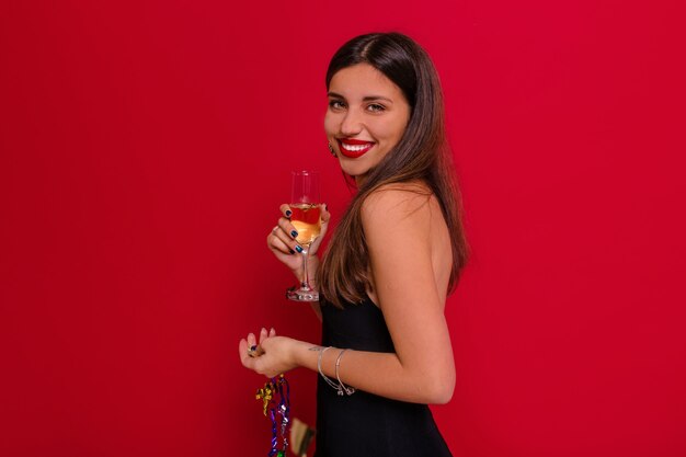 Encantadora dama con gran sonrisa posando sobre pared roja con una copa de champán preparándose para la fiesta de Navidad