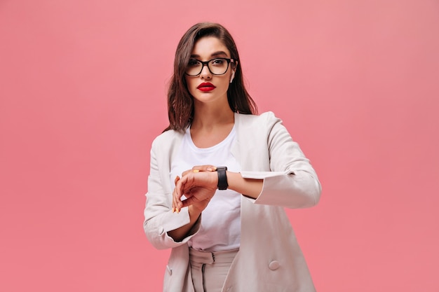 Encantadora dama con labios rojos posando sobre fondo rosa. Grave mujer joven con traje blanco y gafas mira a la cámara sobre fondo aislado.