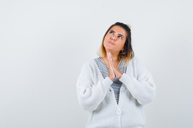 Encantadora dama mostrando las manos entrelazadas en gesto de oración en camiseta, cárdigan y mirando esperanzado