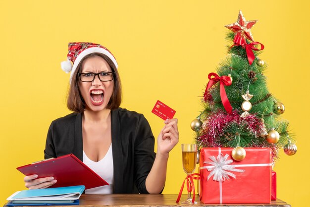 Encantadora dama nerviosa en traje con sombrero de santa claus y anteojos mostrando tarjeta bancaria en la oficina