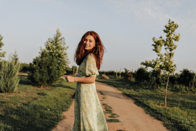 Encantadora dama con peinado astuto en vestido amarillo y verde de moda mirando al frente y sonriendo en la pared de la carretera