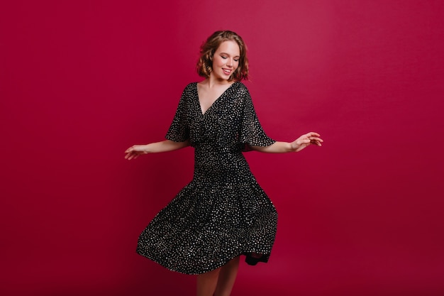 Encantadora dama con pelo corto y brillante bailando en vestido negro vintage y mirando hacia abajo