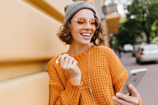 Encantadora dama de pelo corto con sombrero gris de pie junto a la pared con smartphone