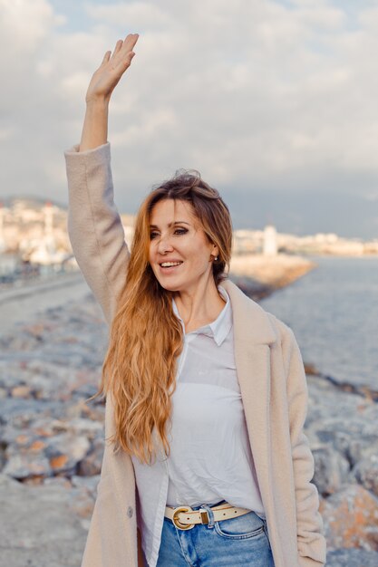 Encantadora dama de pie y agitando su mano en la playa y mirando emocionado durante el día.