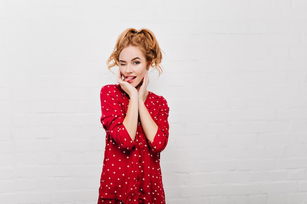 Encantadora dama en pijama rojo haciendo muecas. Retrato de interior de modelo de mujer rubia en traje de noche aislado en la pared blanca.