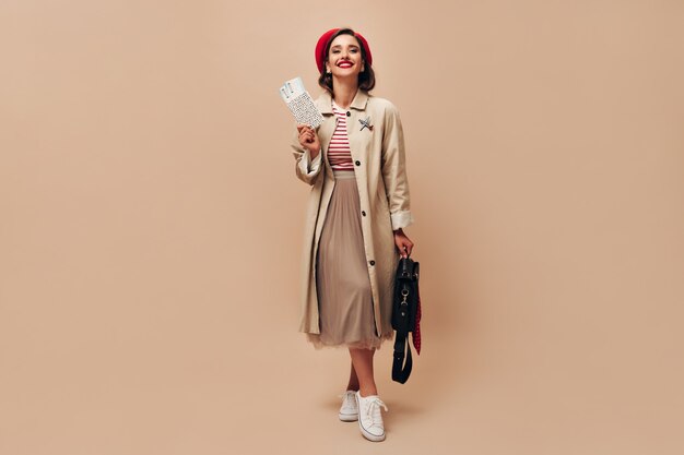 Encantadora dama con sombrero rojo y trinchera beige tiene boletos. Hermosa mujer en elegante abrigo largo y suéter de rayas con bolso negro posando.