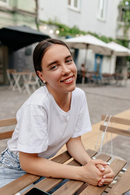 Encantadora dama sonriente con cabello oscuro recogido con camiseta blanca posa en la cámara con una sonrisa maravillosa mientras descansa al aire libre en la cafetería