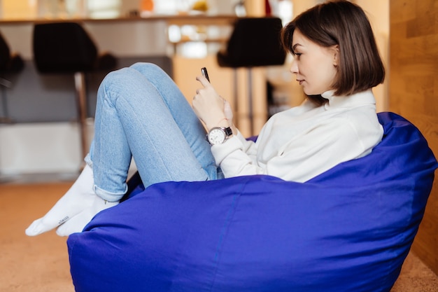 La encantadora dama sonriente está sentada en una silla de bolso violeta brillante usando su teléfono para enviar mensajes de texto con sus amigos