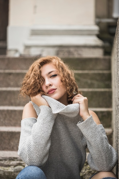 Foto gratuita encantadora dama sonriente sosteniendo cuello de suéter y sentado en pasos