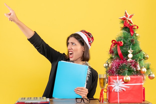 Encantadora dama en traje con sombrero de santa claus y decoraciones de año nuevo con documento apuntando hacia arriba en la oficina en amarillo aislado