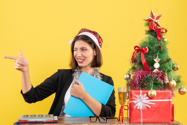 Encantadora dama en traje con sombrero de santa claus y decoraciones de año nuevo con documento haciendo reacciones divertidas en la oficina en amarillo aislado