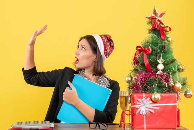 Encantadora dama en traje con sombrero de santa claus y decoraciones de año nuevo con documento mirando su mano en la oficina en amarillo aislado