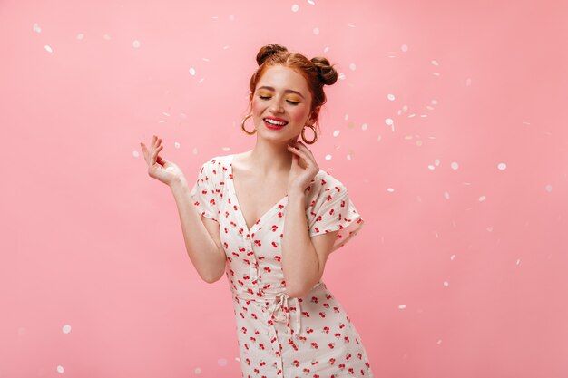 Encantadora dama con vestido blanco con cerezas sonríe afablemente. Retrato de mujer pelirroja en enormes pendientes sobre fondo rosa.