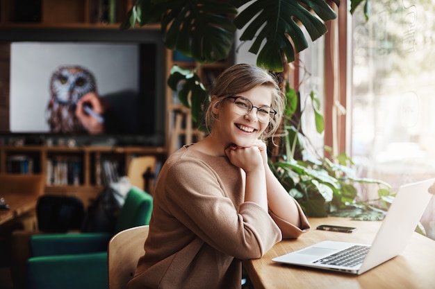 Encantadora y despreocupada mujer europea con cabello rubio con gafas, sentada junto a la ventana y la computadora portátil, apoyándose en las manos.