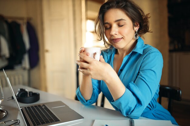 Encantadora y elegante redactora joven que cierra los ojos con placer, disfruta del olor a café de la taza, trabaja en la computadora portátil, escribe un nuevo artículo para las cuentas de las redes sociales, se sienta en su lugar de trabajo