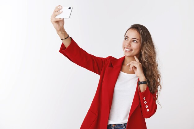 Encantadora empleada de oficina moderna tomando selfie publicando blog en línea diciéndole a los seguidores nuevo trabajo, extendiendo la mano sosteniendo el teléfono inteligente fotografiándose a sí misma sonriendo pantalla de visualización