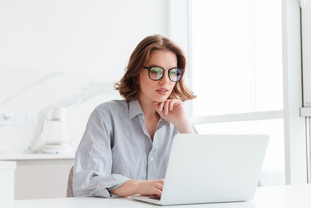 Encantadora empresaria en gafas y camisa a rayas trabajando con la computadora portátil mientras se encuentra en casa