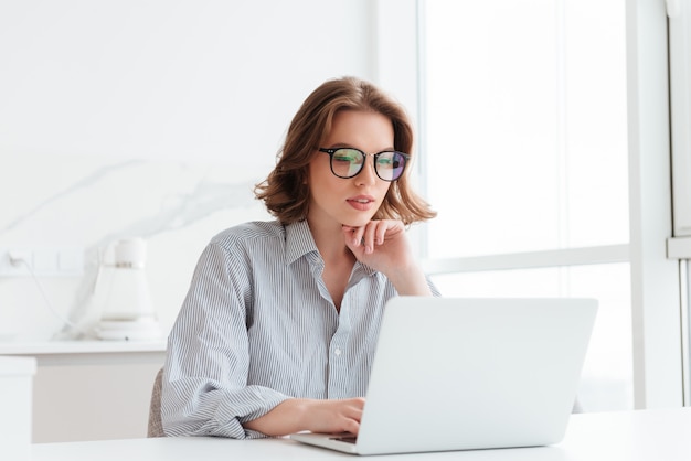 Encantadora empresaria en gafas y camisa a rayas trabajando con la computadora portátil mientras se encuentra en casa