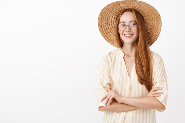 Encantadora exploradora sociable y creativa en blusa de rayas amarillas, sombrero de paja y gafas de moda, cogidos de la mano cruzados sobre el pecho y sonriendo con expresión despreocupada y relajada