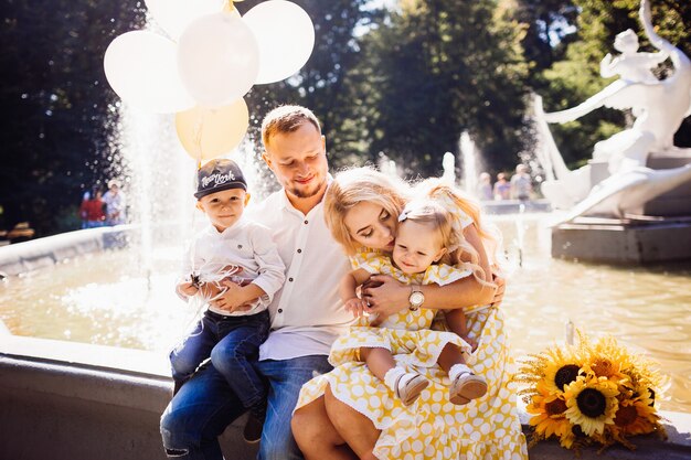 Encantadora familia vestida con la misma ropa sentada en la fuente con sus hijos y globos amarillos