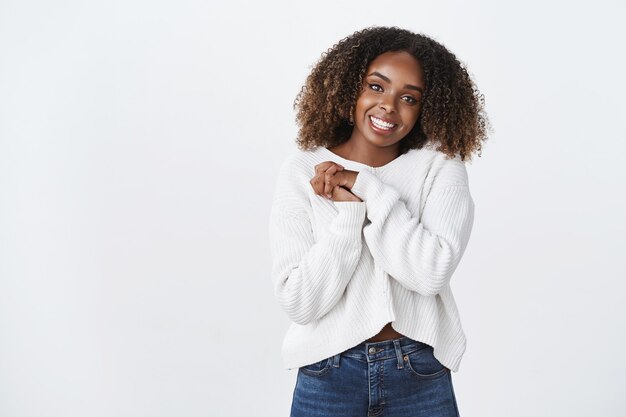 Encantadora feliz sonriente afroamericana de pelo rizado mujer vestida con un suéter blanco presione las palmas juntas gesto de apreciación agradecida, mirando con gratitud la cámara