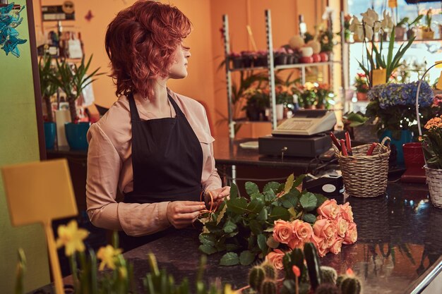 Encantadora floristería pelirroja con uniforme haciendo una hermosa composición floral mientras está de pie en el mostrador de una floristería.